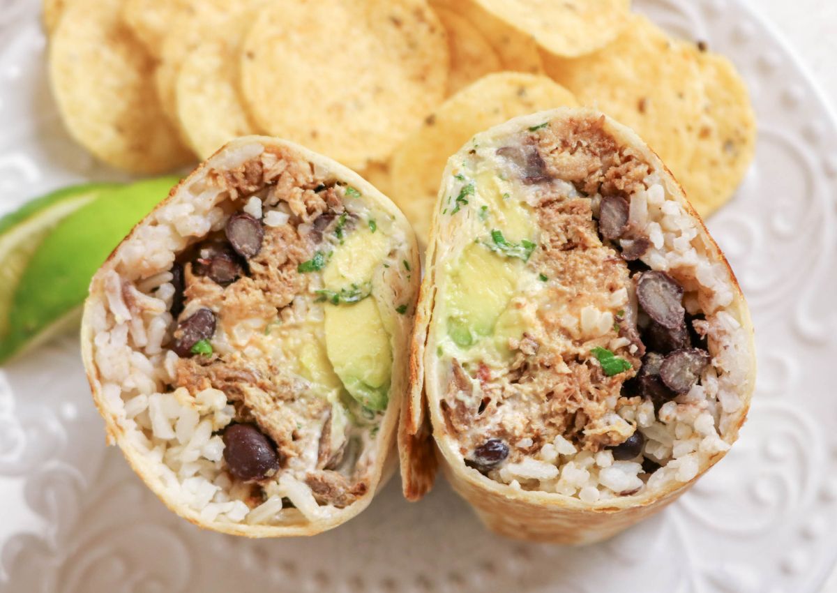 A burrito cut in half shows rice, black beans, shredded meat, avocado, and sauce, with tortilla chips and a lime wedge on a white plate in the background.