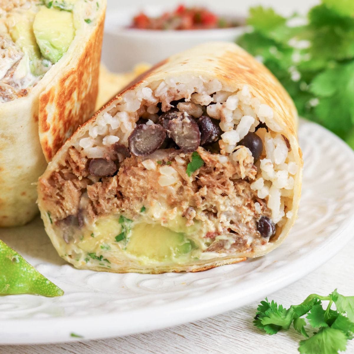 A close-up of a burrito cut in half, showing layers of shredded meat, black beans, rice, avocado, and sauce on a white plate with cilantro and lime.