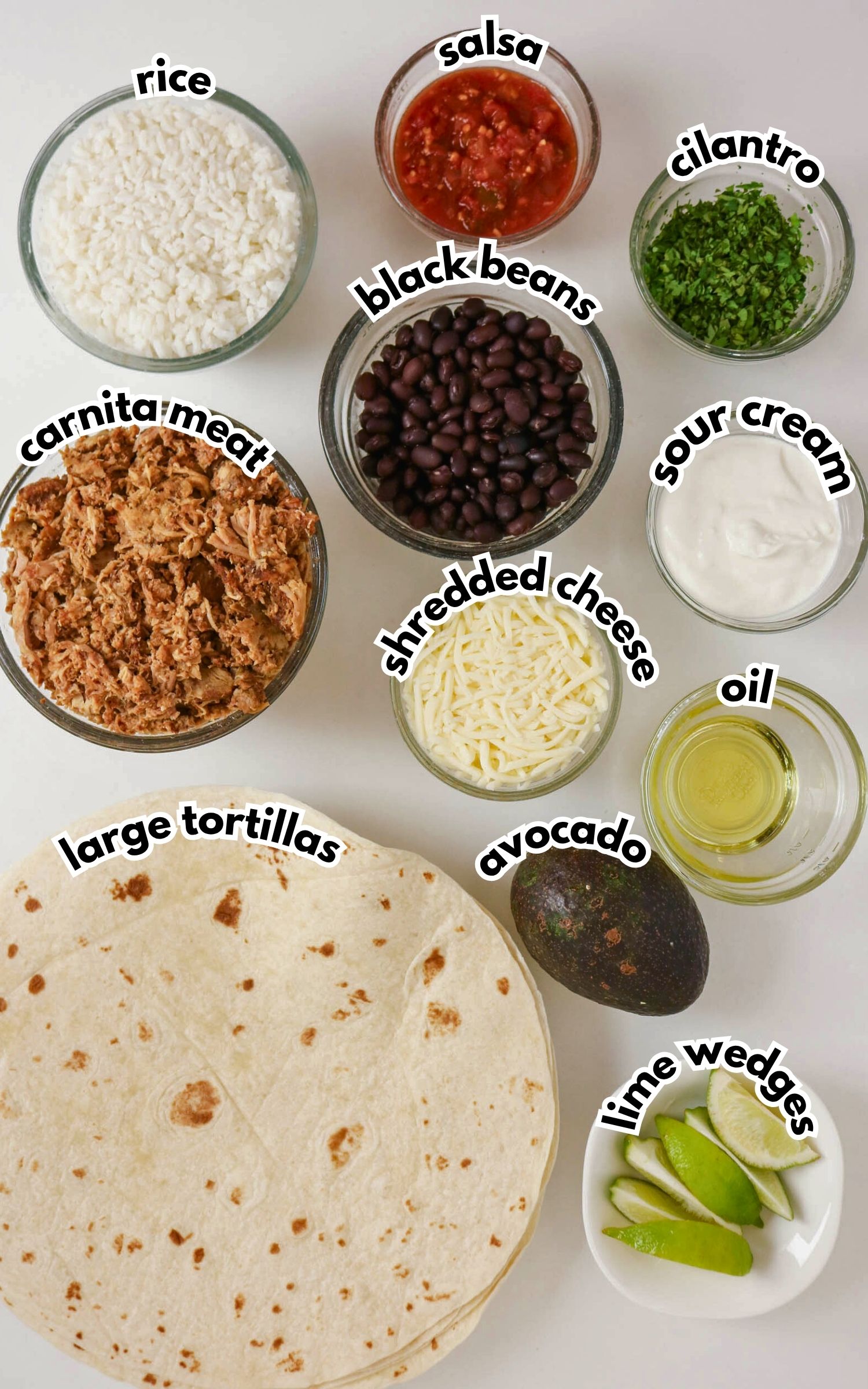 Various burrito ingredients in bowls and on a surface, including rice, carnita meat, large tortillas, black beans, shredded cheese, salsa, cilantro, sour cream, oil, avocado, and lime wedges.