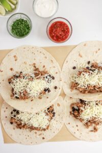 Four flour tortillas topped with seasoned ground meat, black beans, and shredded cheese on parchment paper, with bowls of salsa, sour cream, chopped herbs, and lime wedges nearby.