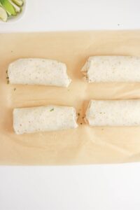 Four wrapped burritos are arranged in a rectangular pattern on a sheet of parchment paper, with lime wedges partially visible in the top left corner.