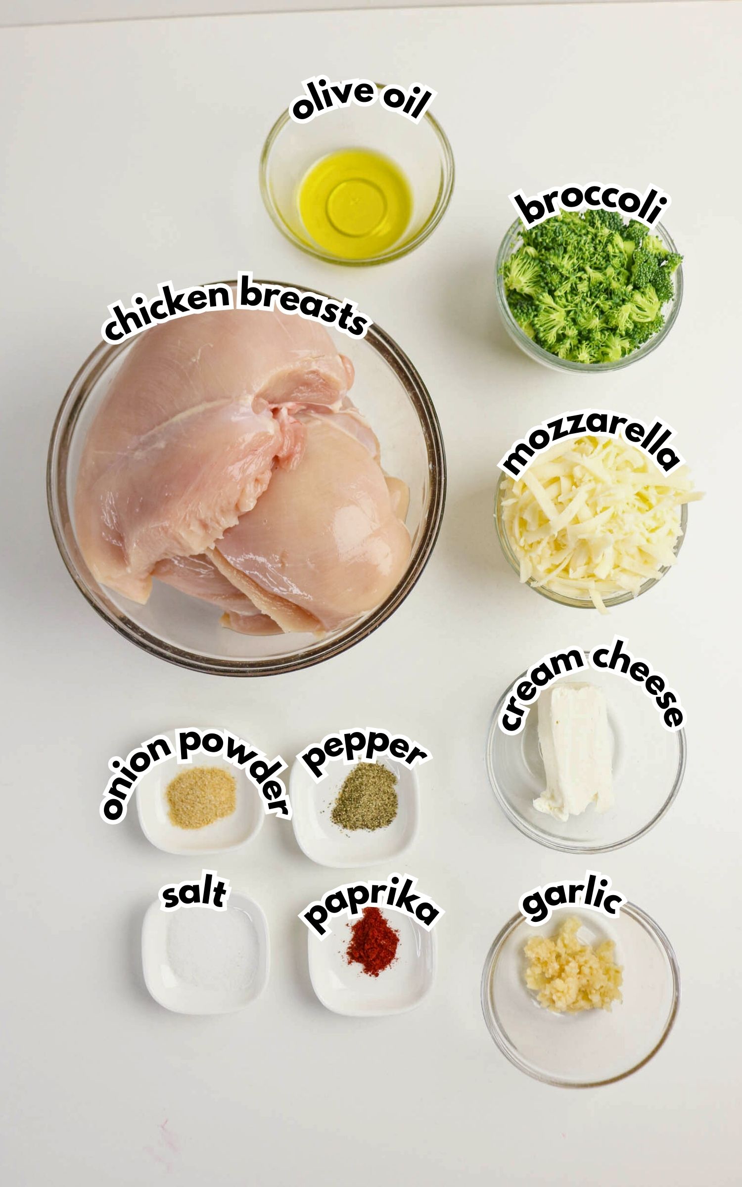Top-down view of separated ingredients: raw chicken breasts, olive oil, chopped broccoli, shredded mozzarella, cream cheese, minced garlic, paprika, pepper, onion powder, and salt in bowls.