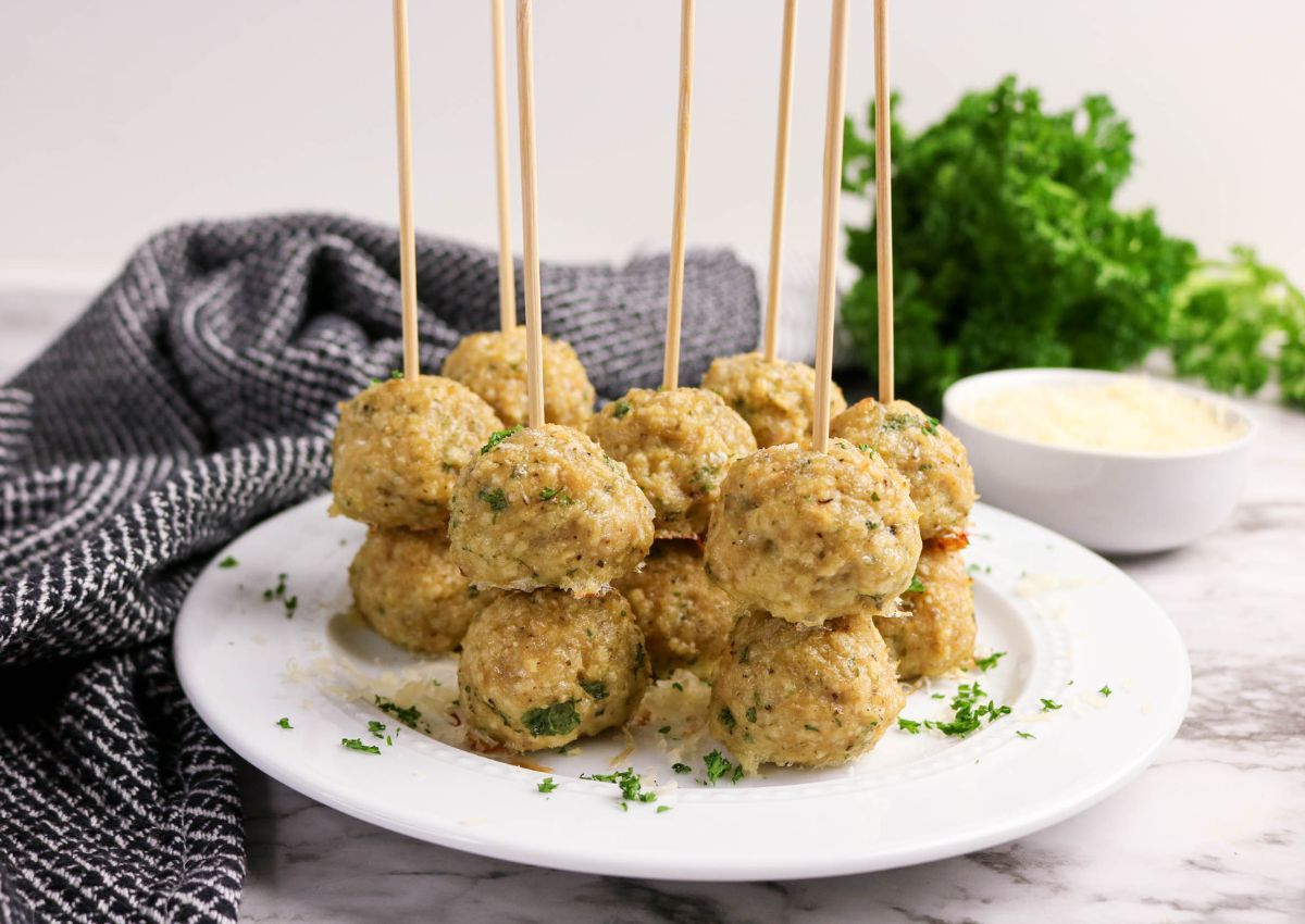 A white plate with skewered meatballs garnished with herbs, placed on a marble surface near a bowl of sauce and fresh parsley.