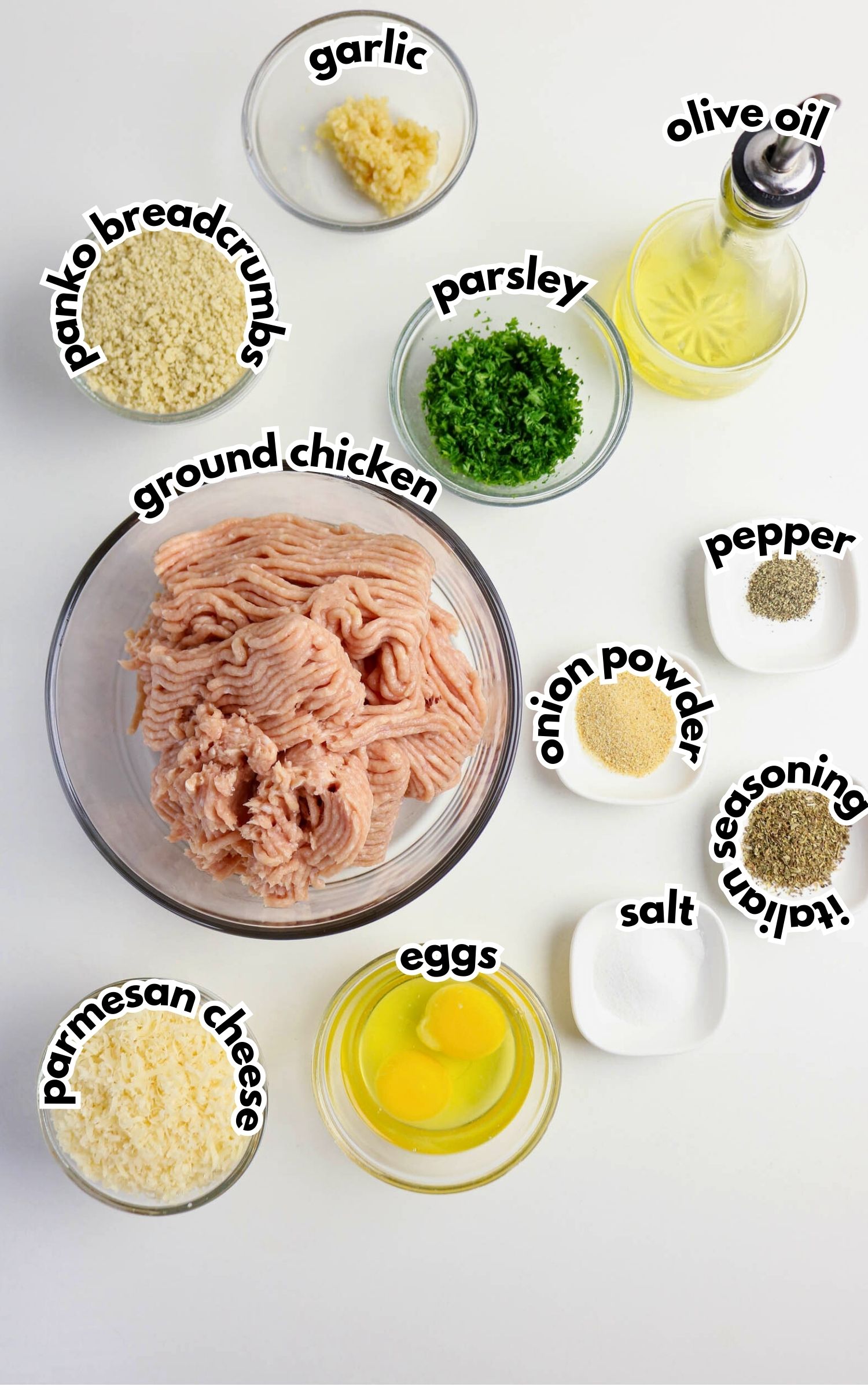 Ingredients for chicken meatballs arranged on a white surface, including ground chicken, eggs, breadcrumbs, parmesan, parsley, garlic, olive oil, and various seasonings.