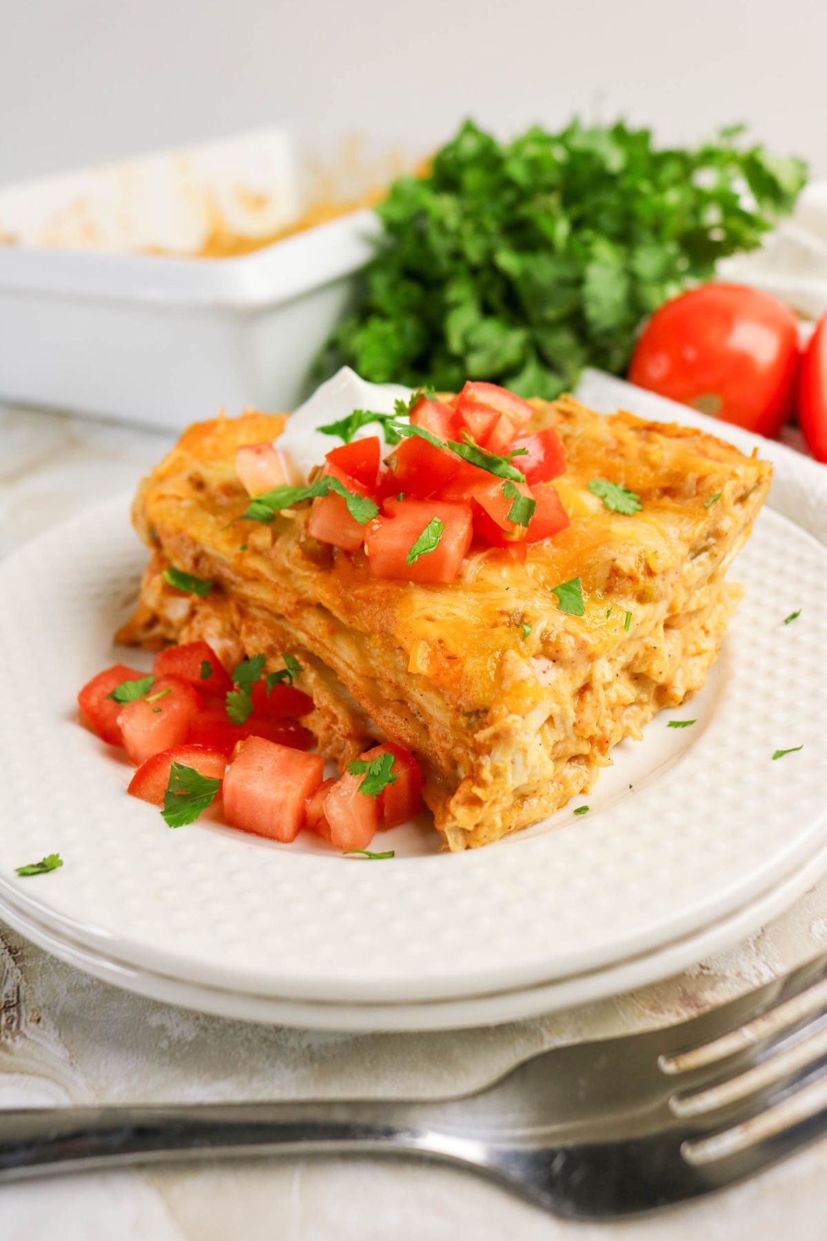 A slice of layered casserole topped with diced tomatoes, cilantro, and sour cream on a white plate, with tomatoes and cilantro in the background.