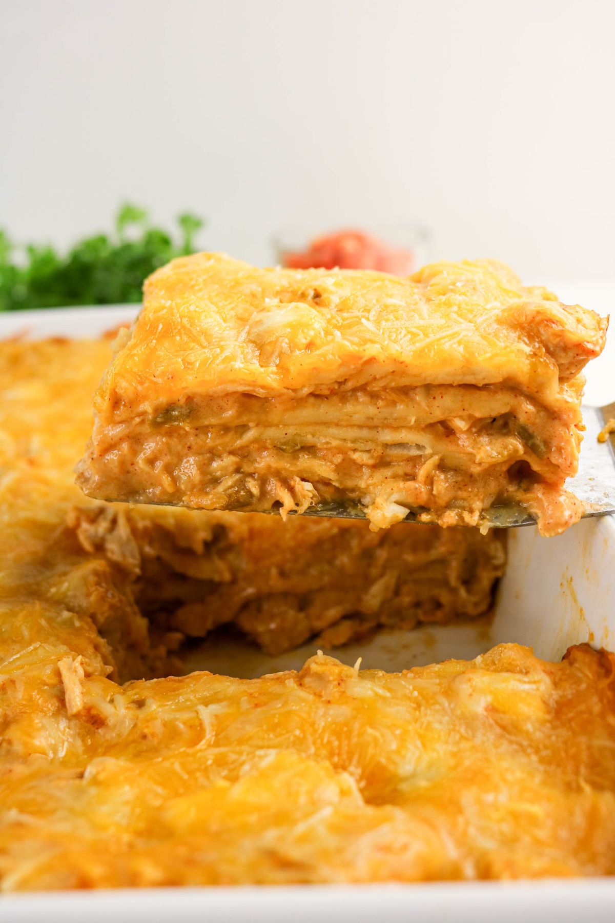 A close-up of a cheesy, golden-brown layered casserole being lifted from a baking dish with a spatula.