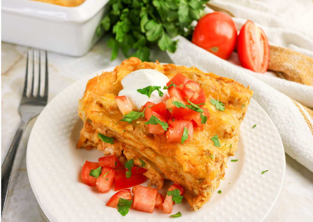 A slice of cheesy lasagna topped with sour cream, diced tomatoes, and cilantro on a white plate, with fresh tomatoes and herbs in the background.