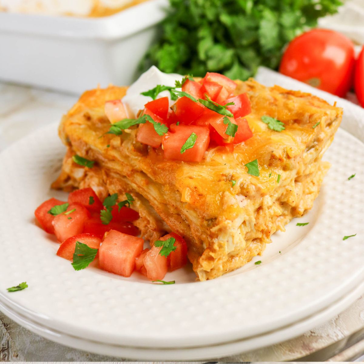 A slice of layered casserole topped with chopped tomatoes, cilantro, and a dollop of sour cream, served on a white plate with fresh tomatoes and greens in the background.