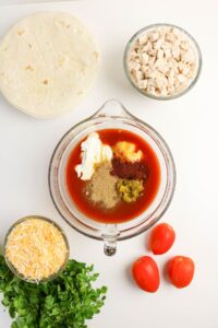 Ingredients for a Mexican dish: tortillas, chopped chicken, shredded cheese, cilantro, tomatoes, and a glass bowl with tomato sauce, sour cream, green chiles, and seasonings.