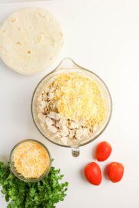 A bowl of diced chicken topped with shredded cheese sits on a white surface next to tortillas, a small bowl of cheese, fresh cilantro, and three Roma tomatoes.