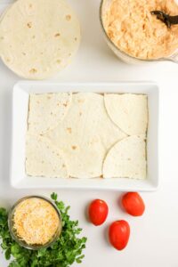 A white dish with tortilla pieces arranged in a single layer, surrounded by tortillas, shredded cheese, tomatoes, chopped herbs, and a bowl of creamy filling on a white surface.