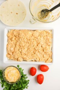 A baking dish with a creamy mixture spread over tortillas, surrounded by shredded cheese, tomatoes, cilantro, extra tortillas, and a mixing bowl with a spoon.