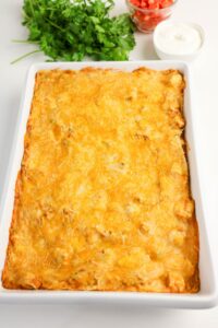 A rectangular casserole dish filled with a baked, cheesy dish sits on a white surface with cilantro, diced tomatoes, and a bowl of sour cream in the background.