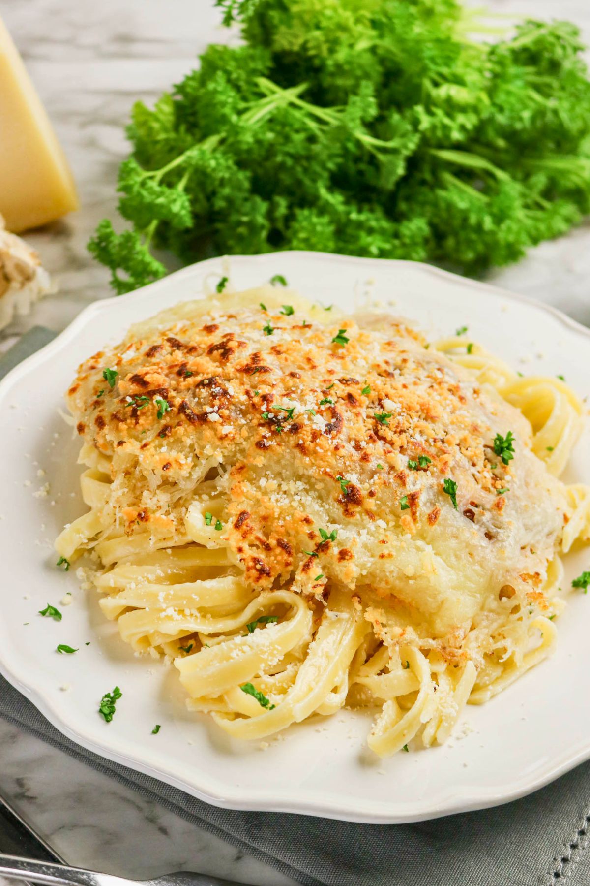 A plate of fettuccine pasta topped with a breaded, baked piece of chicken, garnished with chopped parsley, sits on a white plate with fresh parsley in the background.