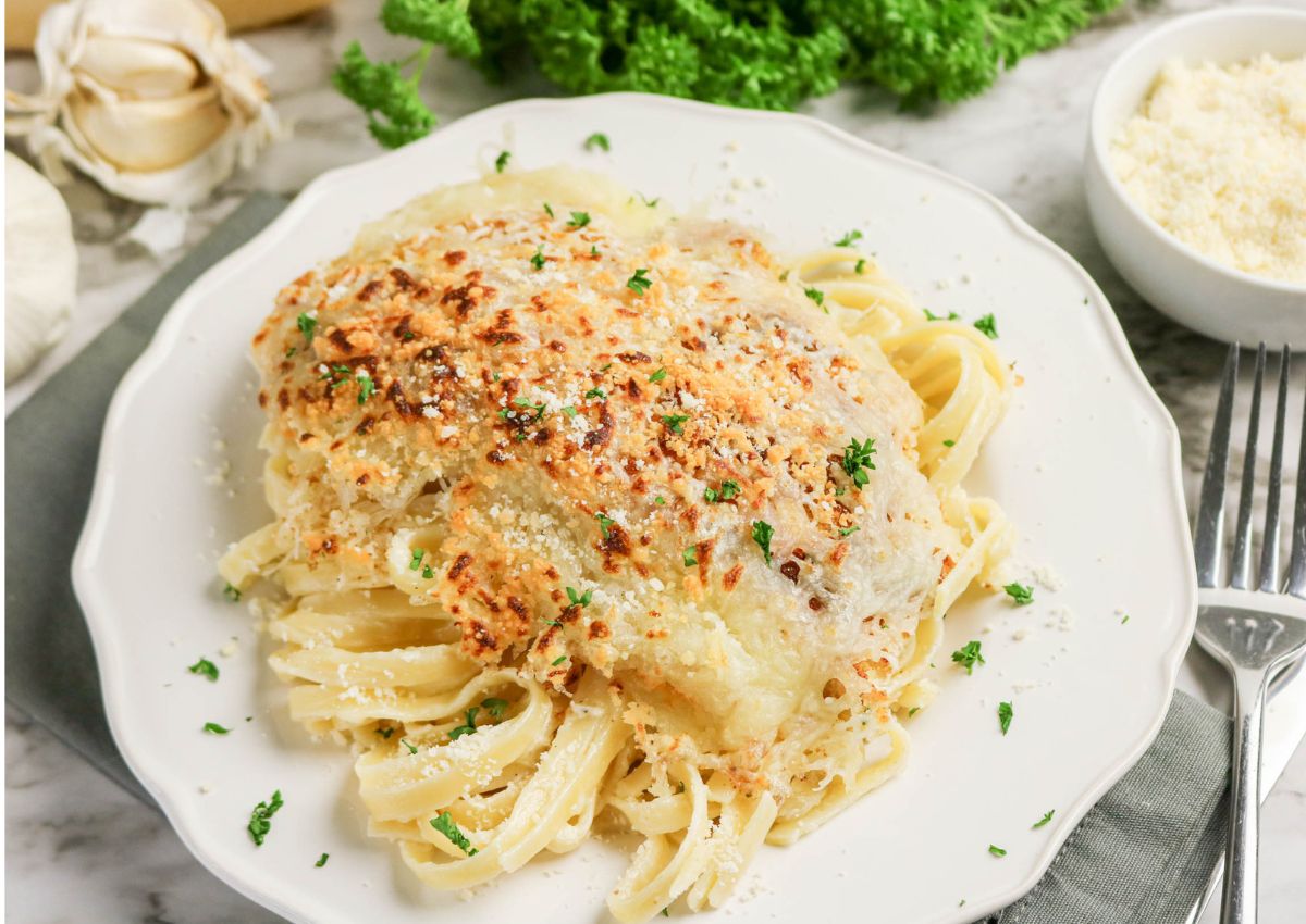 A plate of pasta topped with a baked, cheese-crusted white fish fillet, garnished with chopped parsley, on a white plate beside a fork and a bowl of grated cheese.