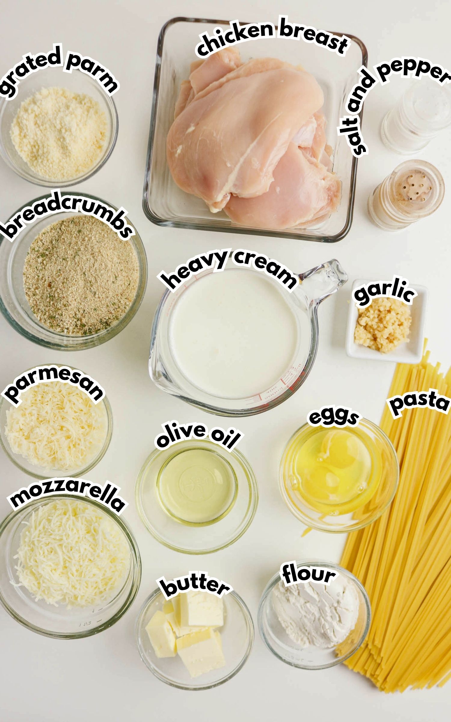 Various ingredients for a pasta dish are arranged on a surface, including raw chicken breast, pasta, cheeses, cream, eggs, breadcrumbs, flour, butter, olive oil, garlic, and seasonings.