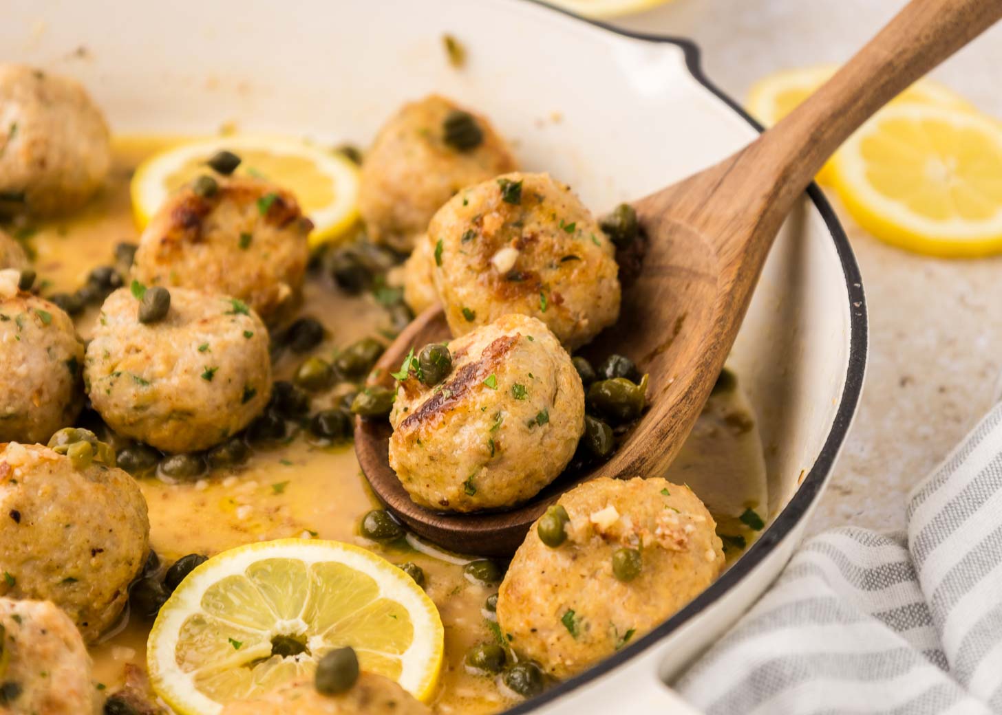 Chicken meatballs with capers and lemon slices in a white skillet, with a wooden spoon lifting two meatballs.