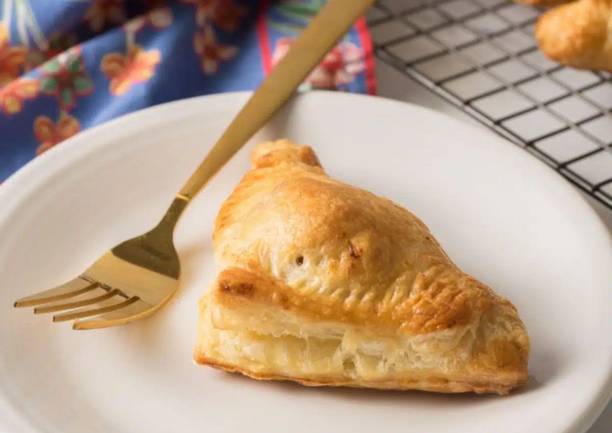 A golden-brown puff pastry turnover sits on a white plate with a gold fork, next to a patterned cloth.
