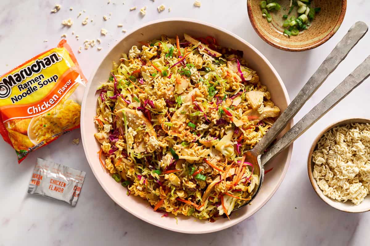 A bowl of ramen noodle salad with shredded vegetables, metal utensils, a packet of Maruchan ramen, seasoning packet, and a dish of chopped green onions nearby.