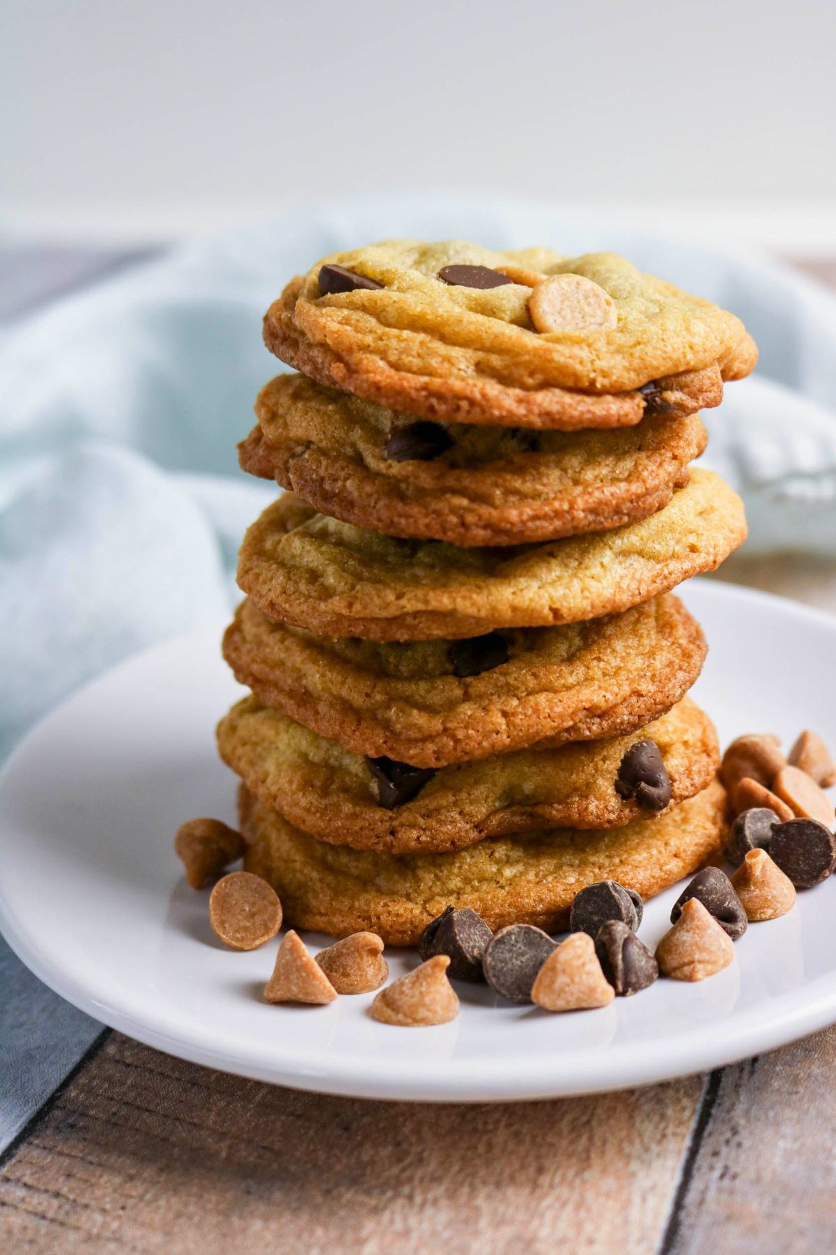 A stack of chocolate chip cookies on a white plate with scattered chocolate and peanut butter chips around them.