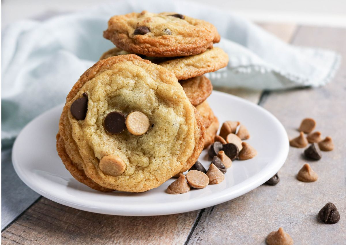 A white plate holds a stack of chocolate chip cookies, with chocolate and peanut butter chips scattered on the plate and wooden surface.