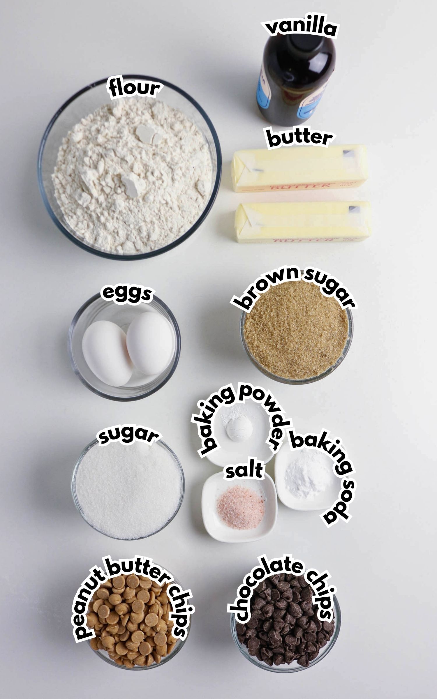 Various baking ingredients are arranged on a white surface, including flour, butter, vanilla, eggs, brown sugar, sugar, baking powder, baking soda, salt, peanut butter chips, and chocolate chips.
