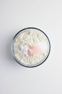 A glass bowl containing a mixture of flour, baking powder, baking soda, and pink salt on a white surface.