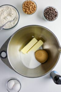 A mixing bowl with butter, white sugar, and brown sugar, surrounded by bowls of flour, chocolate chips, peanut butter chips, eggs, and a bottle of vanilla extract.