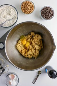 A mixing bowl with cookie dough, eggs, and sugar in the center, surrounded by bowls of flour, peanut butter chips, chocolate chips, vanilla extract, and baking utensils.