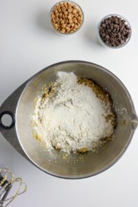 A metal mixing bowl with flour on top of cookie dough, beside bowls of peanut butter chips and chocolate chips, and a hand mixer on a white surface.