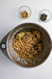 A mixing bowl with cookie dough, peanut butter chips, and chocolate chips, with two small bowls of more chips on a white surface.