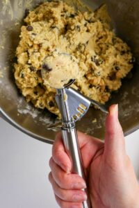 A hand holds a metal cookie scoop with a portion of chocolate chip cookie dough over a mixing bowl filled with more dough.