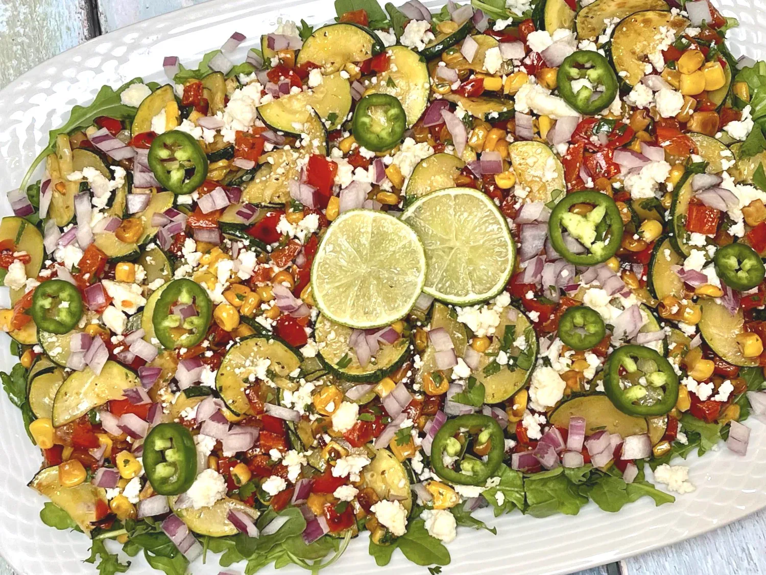 A platter of salad with sliced zucchini, corn, red onion, red bell pepper, jalapeño, crumbled cheese, and lime slices on a bed of greens.