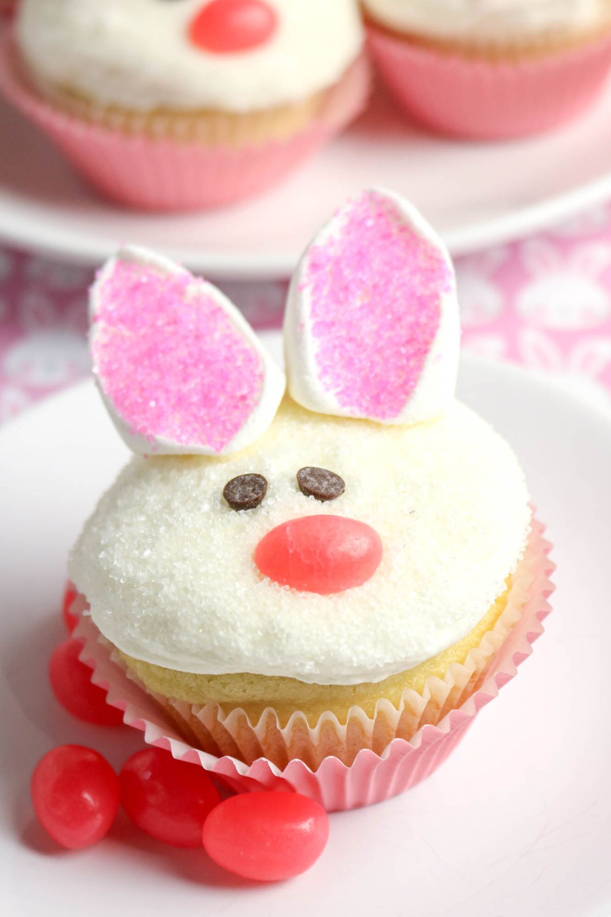 A cupcake decorated to look like a bunny face with marshmallow ears, pink sugar, chocolate chip eyes, a jellybean nose, and surrounded by pink jellybeans.