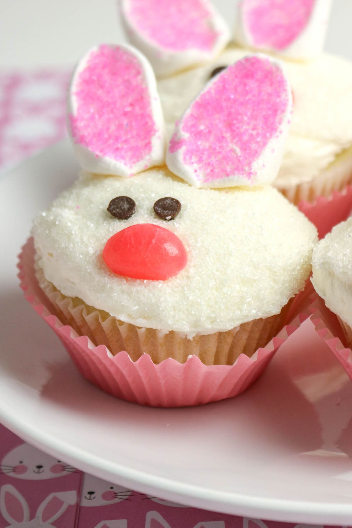 Cupcake decorated as a bunny with pink and white ears, a pink candy nose, and chocolate chip eyes on a white plate.