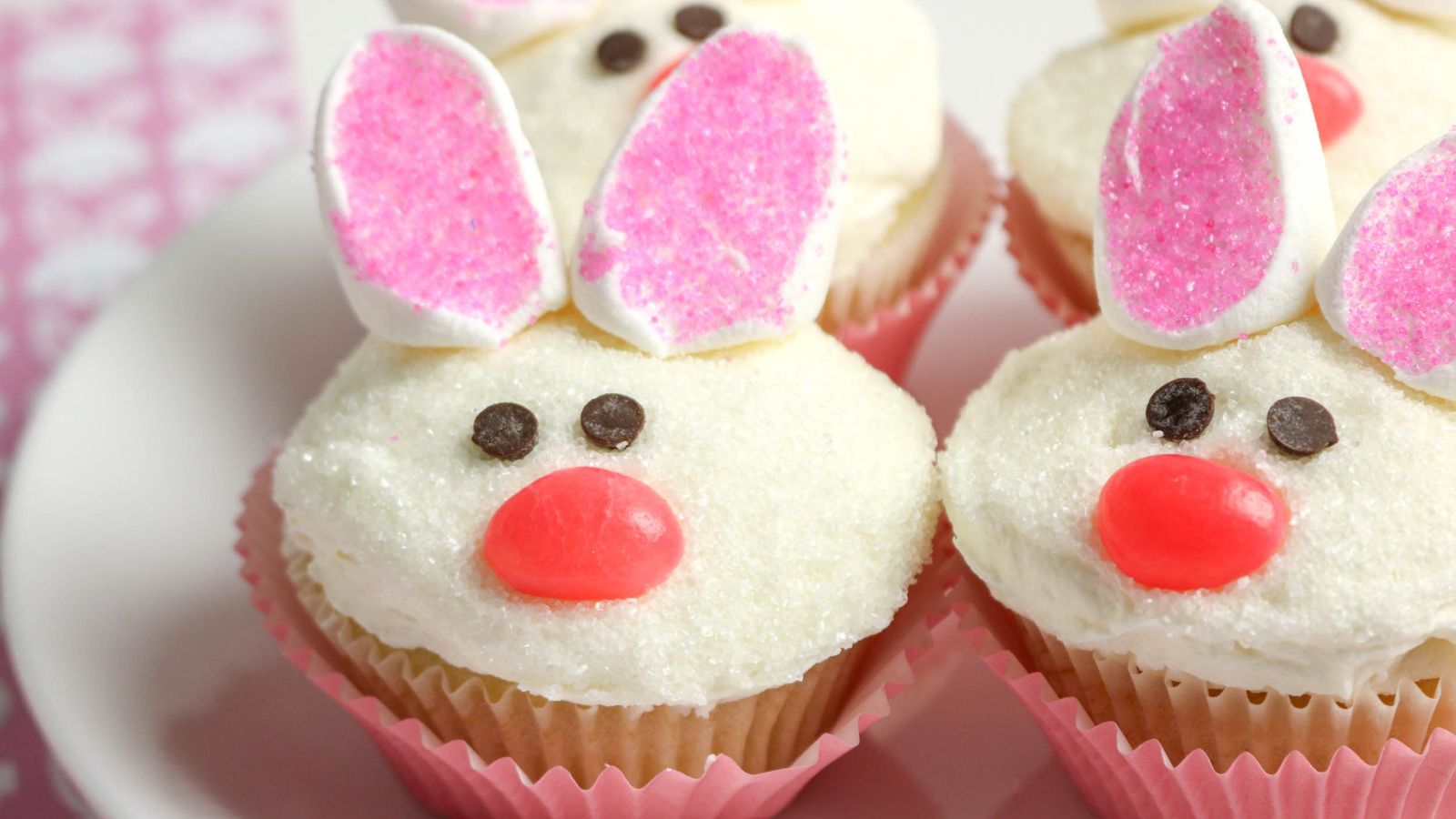 Four cupcakes decorated to look like bunny faces with white frosting, pink marshmallow ears, chocolate chip eyes, and red candy noses, arranged on a white plate.
