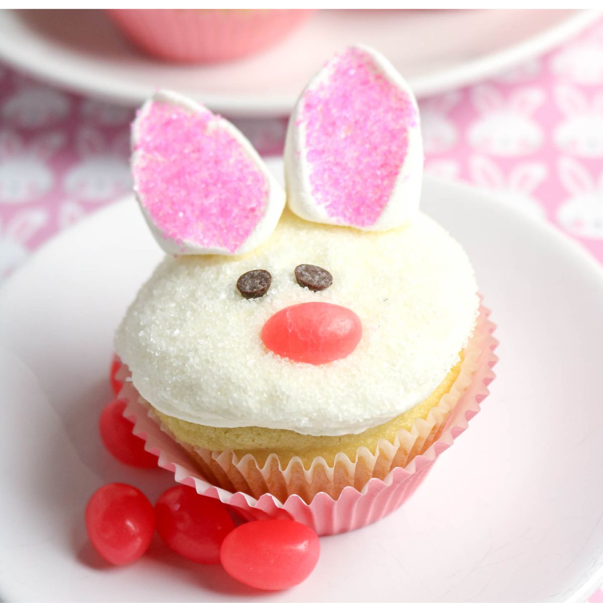 Vanilla cupcake decorated as a bunny with marshmallow ears, chocolate chip eyes, and a pink candy nose, surrounded by pink jelly beans on a white plate.