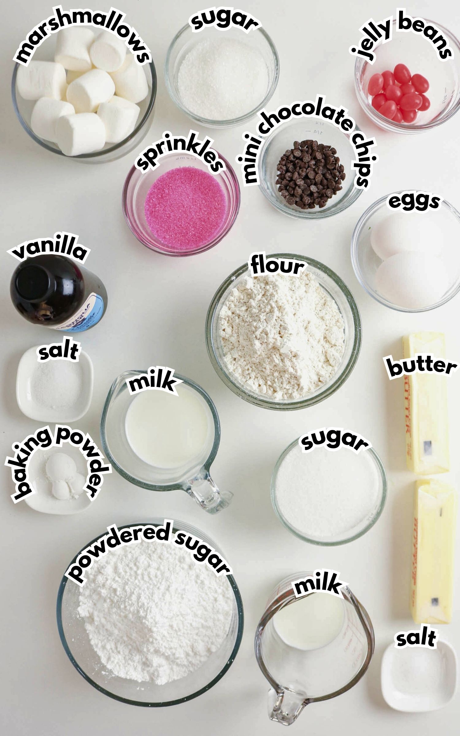 Various baking ingredients are arranged in bowls, including marshmallows, sugar, jelly beans, sprinkles, mini chocolate chips, eggs, vanilla, flour, milk, butter, baking powder, salt, and powdered sugar.