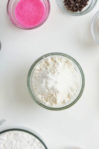 A glass bowl filled with flour sits on a white surface, surrounded by bowls of pink sugar, chocolate chips, and other baking ingredients.