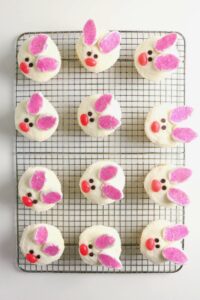 Cupcakes decorated as bunny faces with pink ears, red noses, and chocolate eyes, arranged on a wire cooling rack.