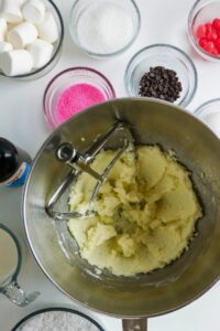 A mixing bowl with creamed butter and sugar, surrounded by ingredients like marshmallows, sugar, pink sprinkles, chocolate chips, vanilla, and milk in glass containers.
