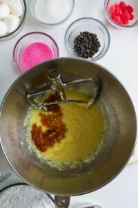 A mixing bowl with creamed ingredients and vanilla extract, surrounded by bowls of sugar, pink sprinkles, chocolate chips, marshmallows, and red candies.