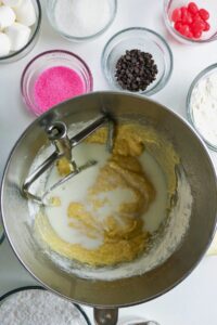 A stand mixer bowl with partially mixed batter, surrounded by bowls of sugar, flour, chocolate chips, marshmallows, pink sprinkles, and red candy.
