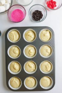 A muffin tray with 12 cupcake liners filled with batter is on a table, surrounded by bowls of pink sugar, mini chocolate chips, marshmallows, shredded coconut, and red candies.