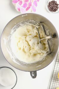 A metal mixing bowl with creamy batter and a paddle attachment, surrounded by sugar, chocolate chips, and pink-topped marshmallows.
