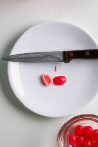 A paring knife and two red jellybeans, one cut in half, are on a white plate with a glass bowl of more jellybeans partially visible at the bottom right.