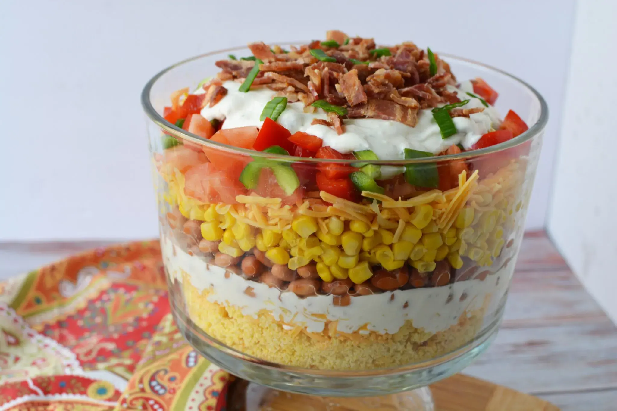 A glass trifle bowl filled with layered salad, including cornbread, beans, corn, cheese, tomatoes, lettuce, sour cream, and bacon bits, displayed on a wooden surface.