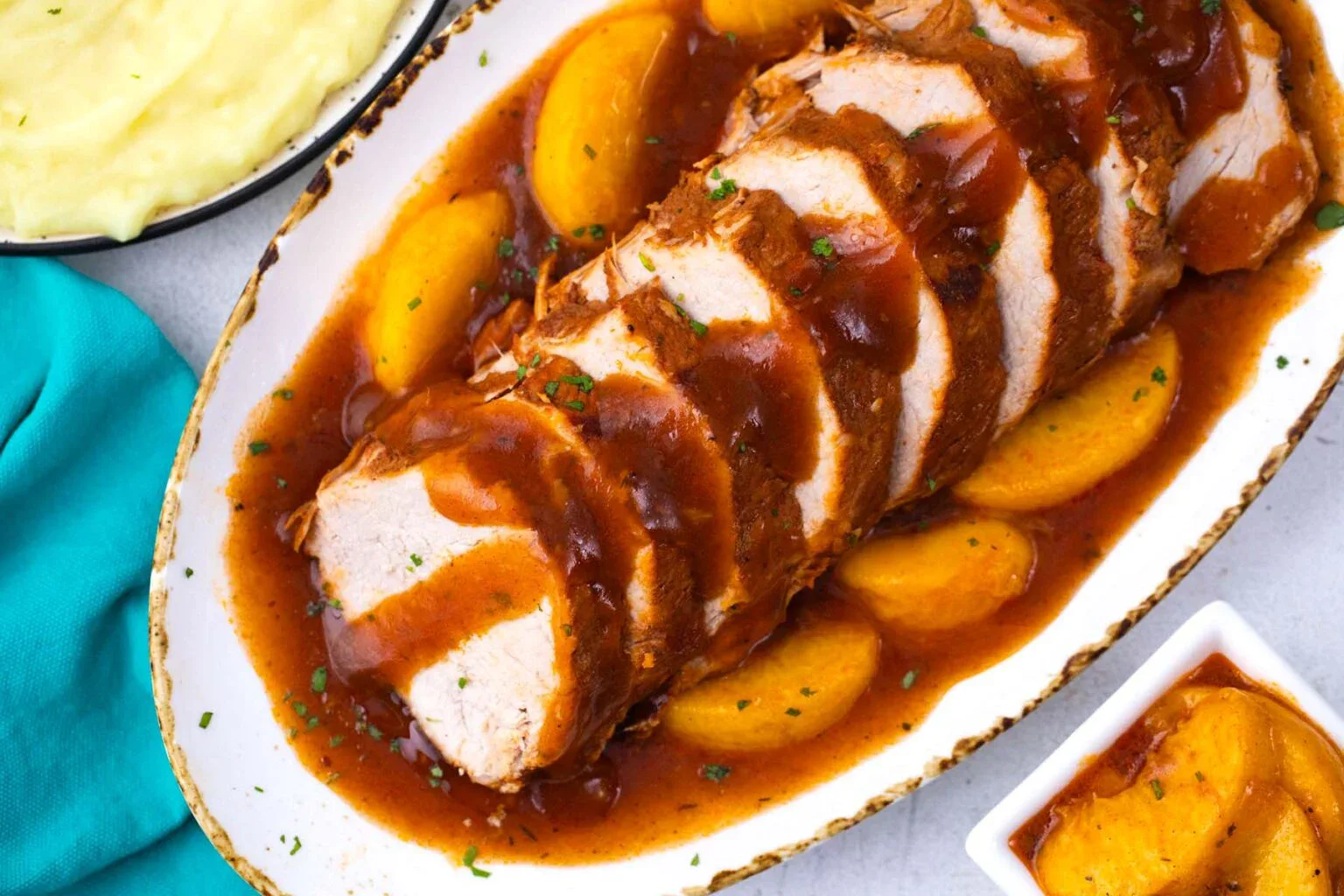 Sliced pork loin with brown sauce and peach slices on an oval plate, served with mashed potatoes and extra peach slices on the side.