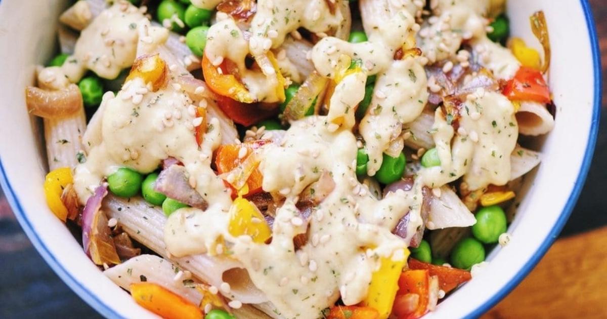 A bowl of penne pasta mixed with peas, bell peppers, onions, and topped with a creamy sauce and sprinkled with herbs and seeds.