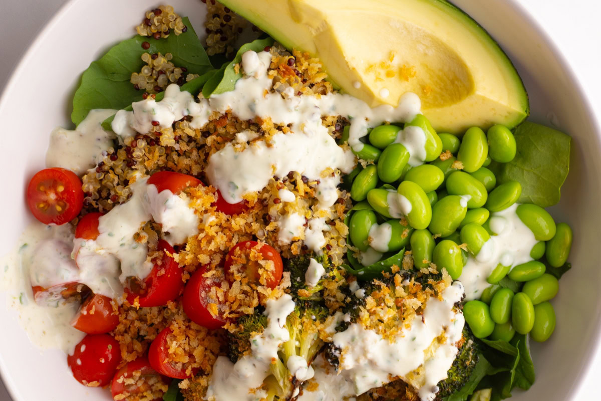A bowl containing sliced avocado, edamame, cherry tomatoes, quinoa, greens, and roasted broccoli, topped with a creamy white dressing and breadcrumbs.
